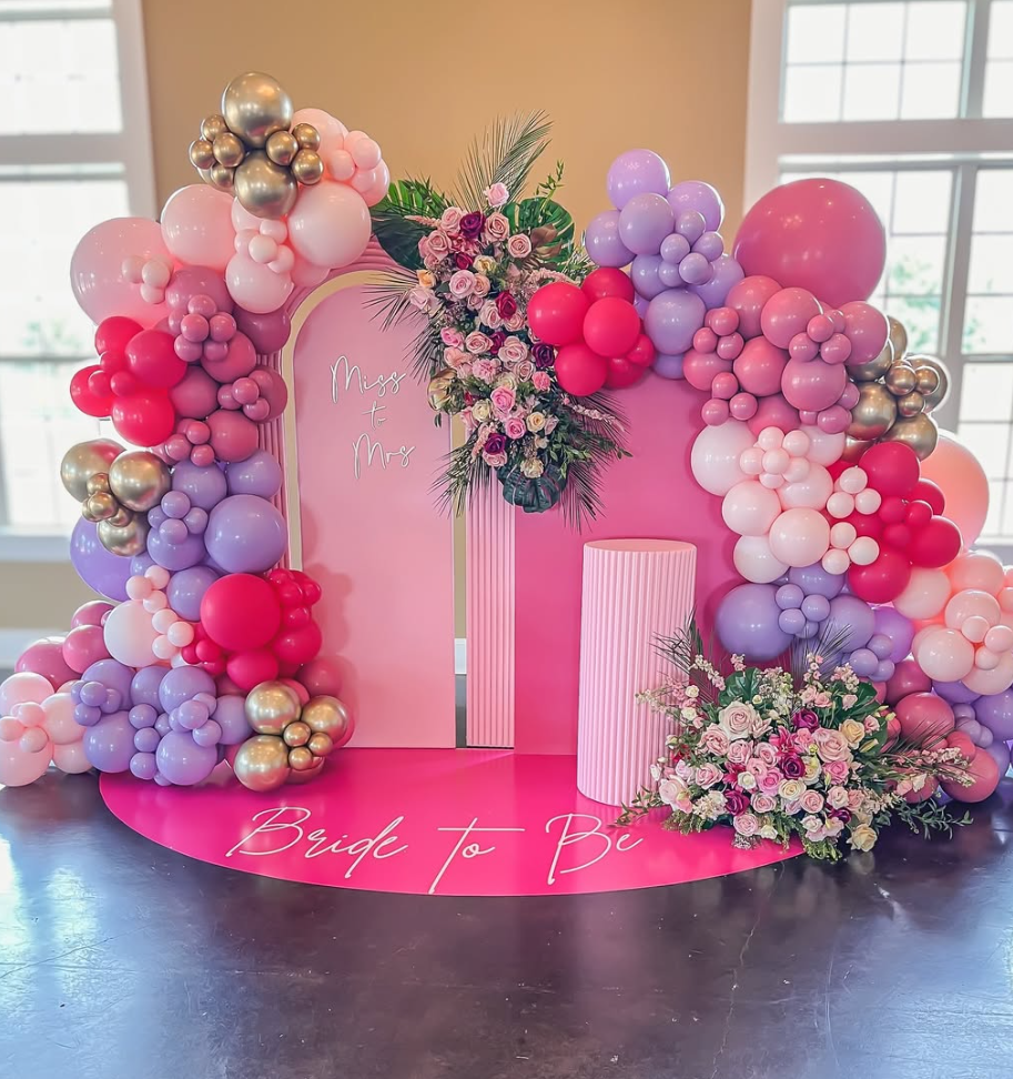 Pink decorative backdrop with balloon arrangements in pink, purple, red, and gold, floral arrangements, and a pink cylindrical pedestal. The backdrop has the words "Miss to Mrs" and the floor says "Bride to Be."