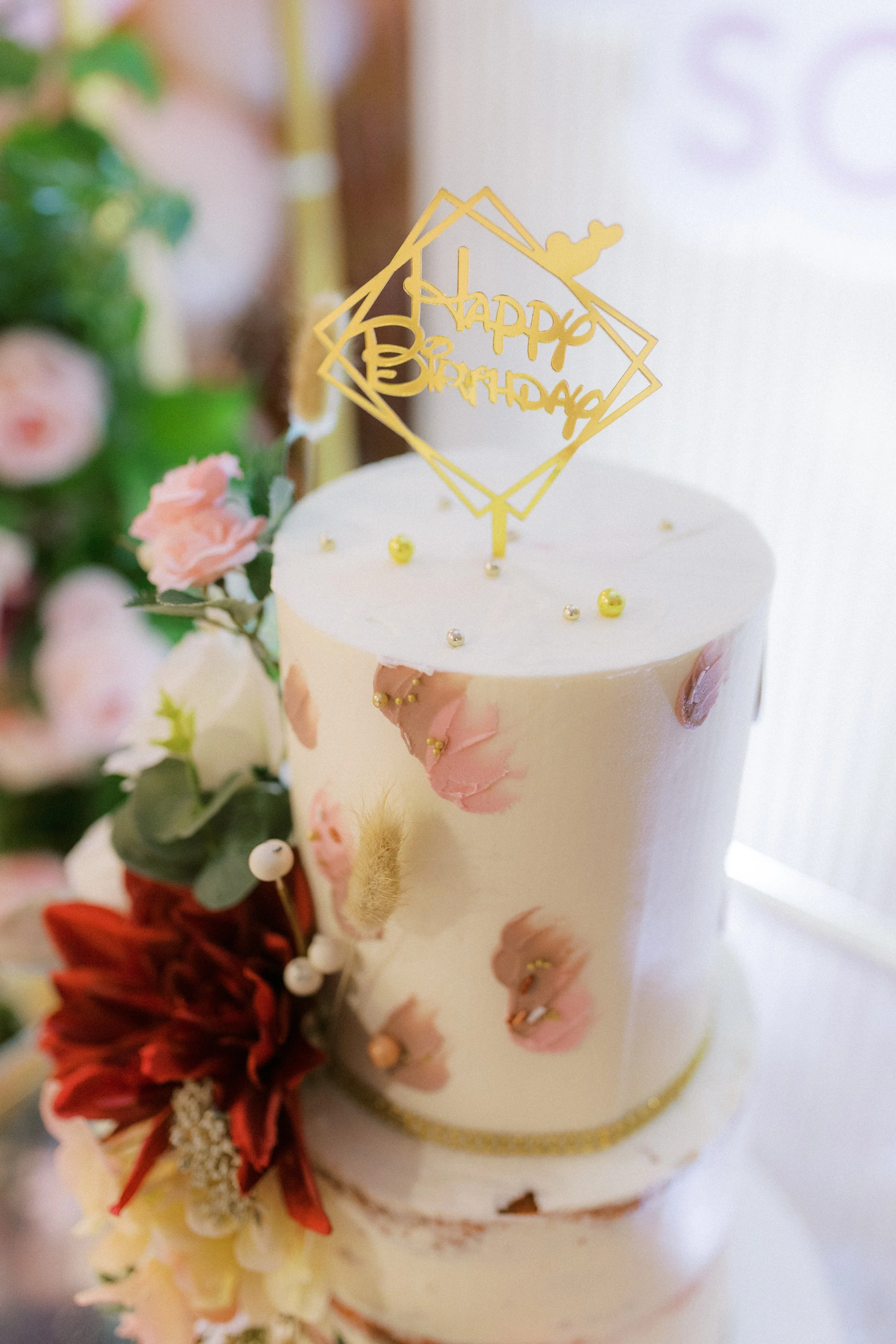 Decorative birthday cake with a gold 'Happy Birthday' topper, pink and peach flowers, and gold and pearl accents.
