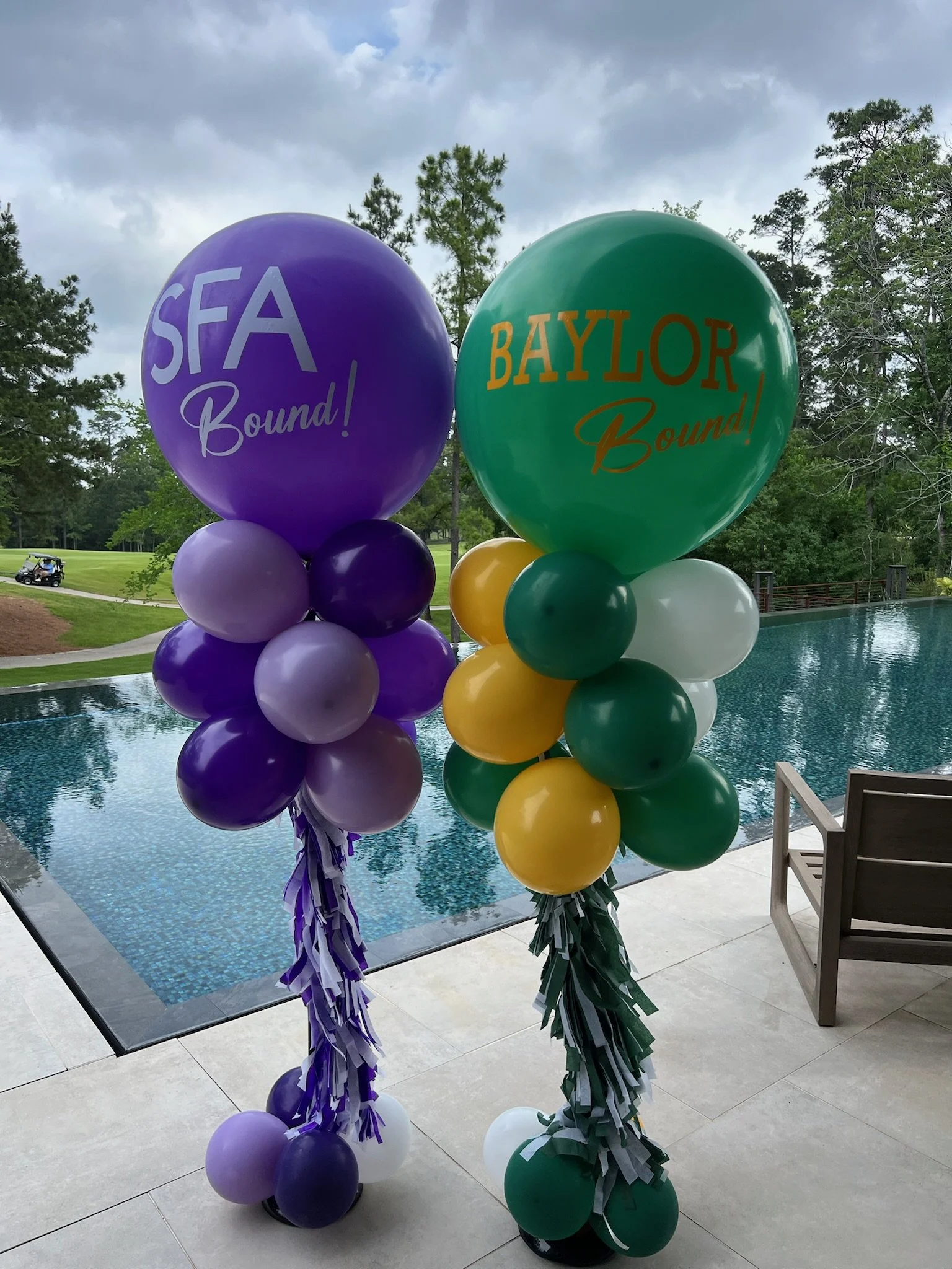 Colorful graduation balloons at a poolside outdoor setting, with trees and cloudy sky in the background.