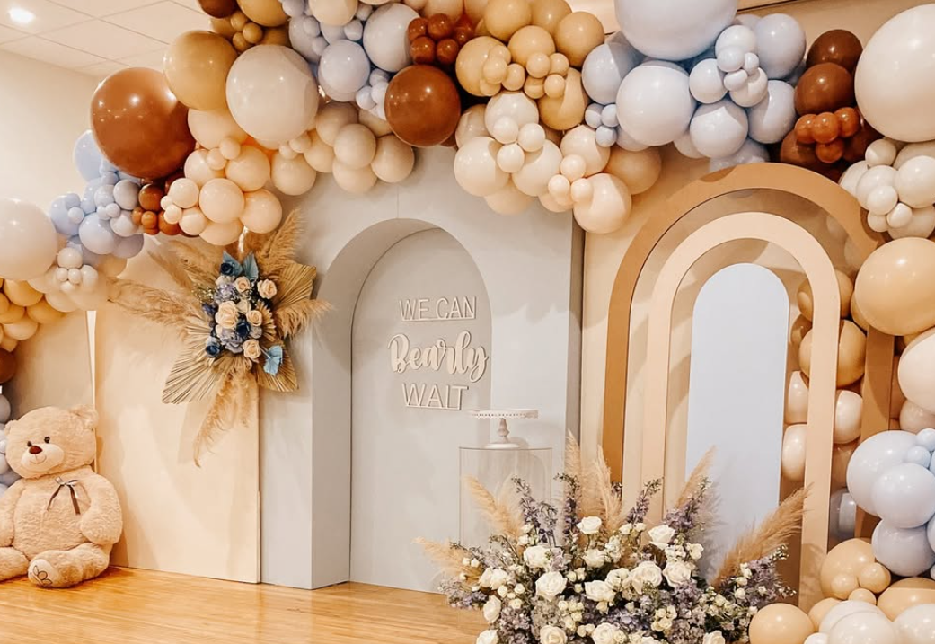 Decorative balloon arch with beige, brown, white, and light blue balloons, cream-colored floral arrangement with pampas grass, teddy bear, and a sign that says "We can really wait" on a pastel wall, with decorative arches.