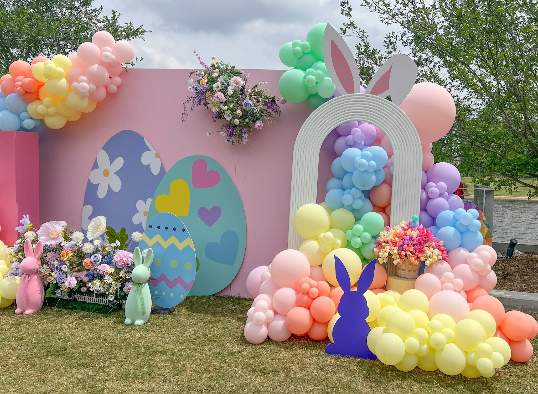 Decorative Easter-themed setup with pastel-colored balloons, large bunny silhouette cutouts, painted egg decorations, and floral arrangements on a pink backdrop outdoors near water.