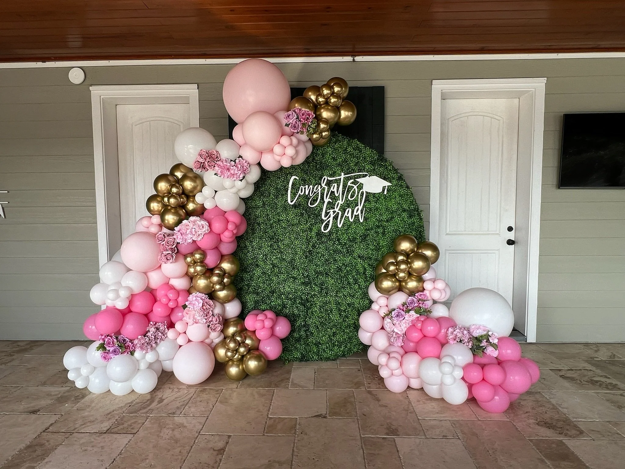 A balloon and floral decoration with pink, white, and gold balloons, and pink flowers, arranged around a green hedge with a white sign that reads "Congrat Grads".