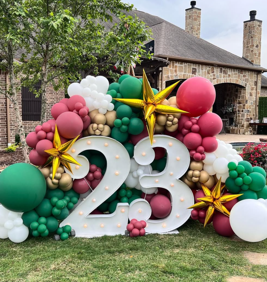 Decorative balloon arrangement with large white '2023' numbers, surrounded by green, red, beige, and white balloons, and gold star-shaped balloons, in front of a house with a stone facade and chimneys, on a grassy lawn.