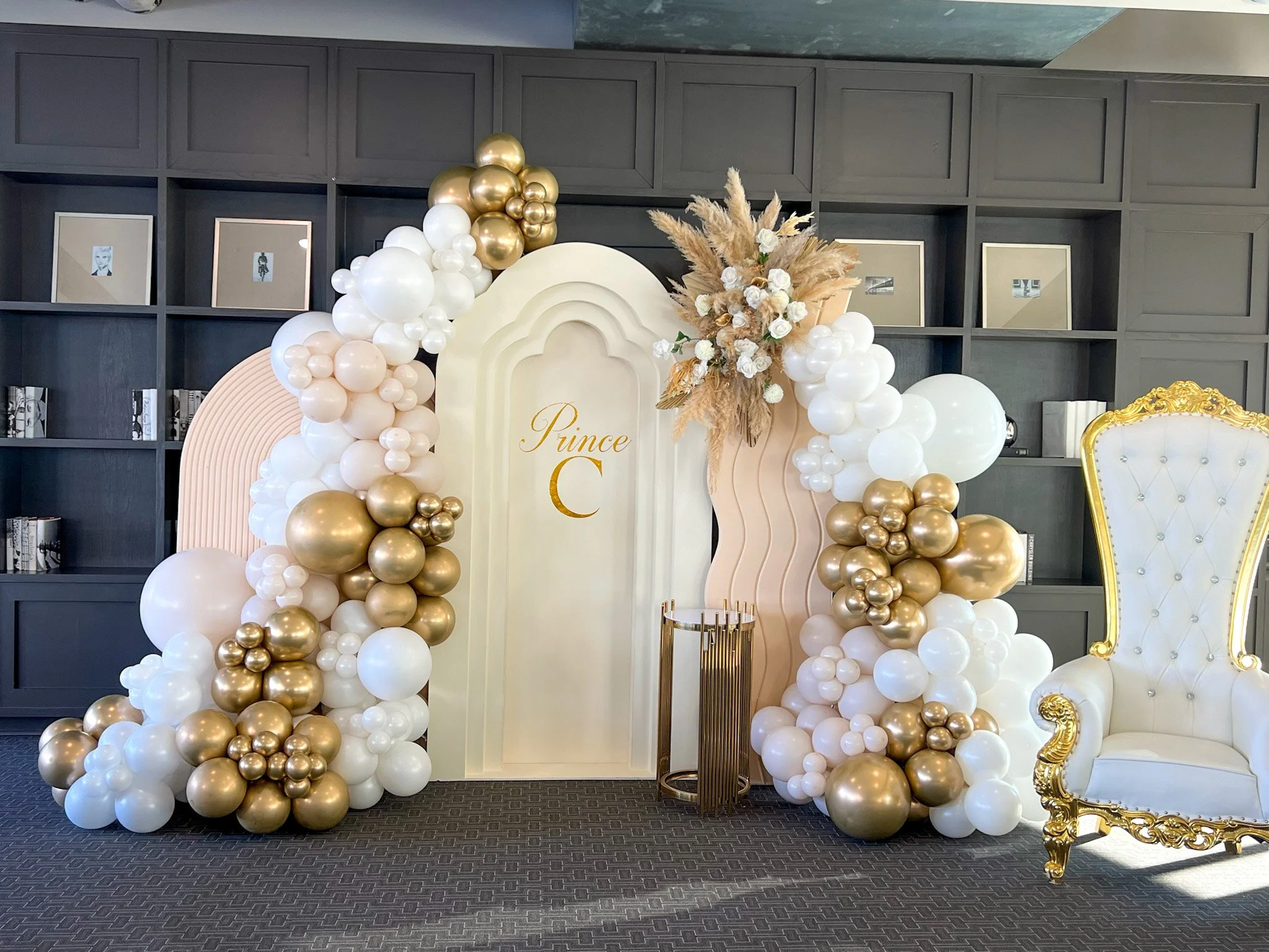 Decorative balloon arch with white, gold, and blush pink balloons, a floral arrangement of white roses and pampas grass, and a backdrop with the name "Prince C" in gold lettering. An elegant white and gold throne-style chair is on the right.