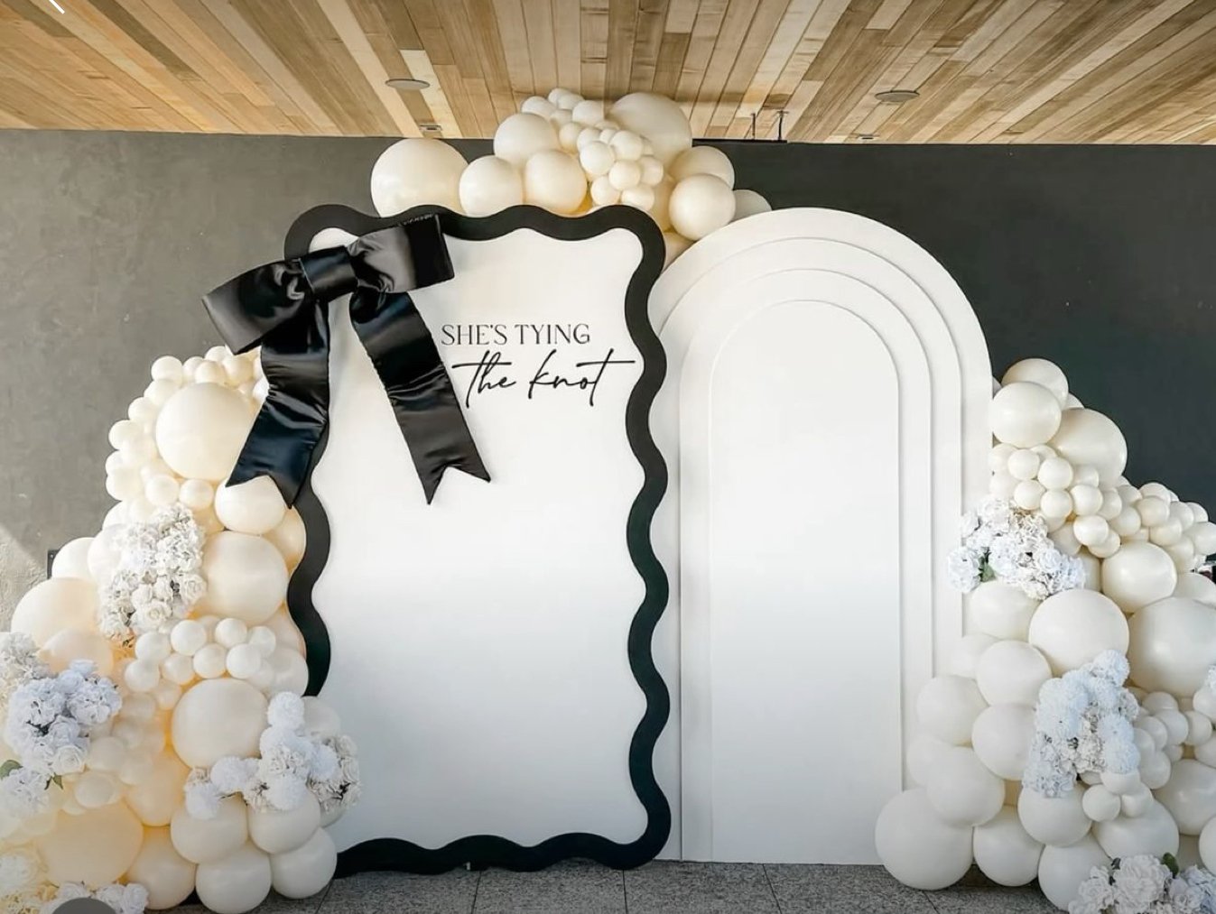 A decorative backdrop with a black scalloped frame, white balloon garland, white flowers, and a black satin bow, with the text "SHE'S TYING the knot" in black cursive and serif fonts.