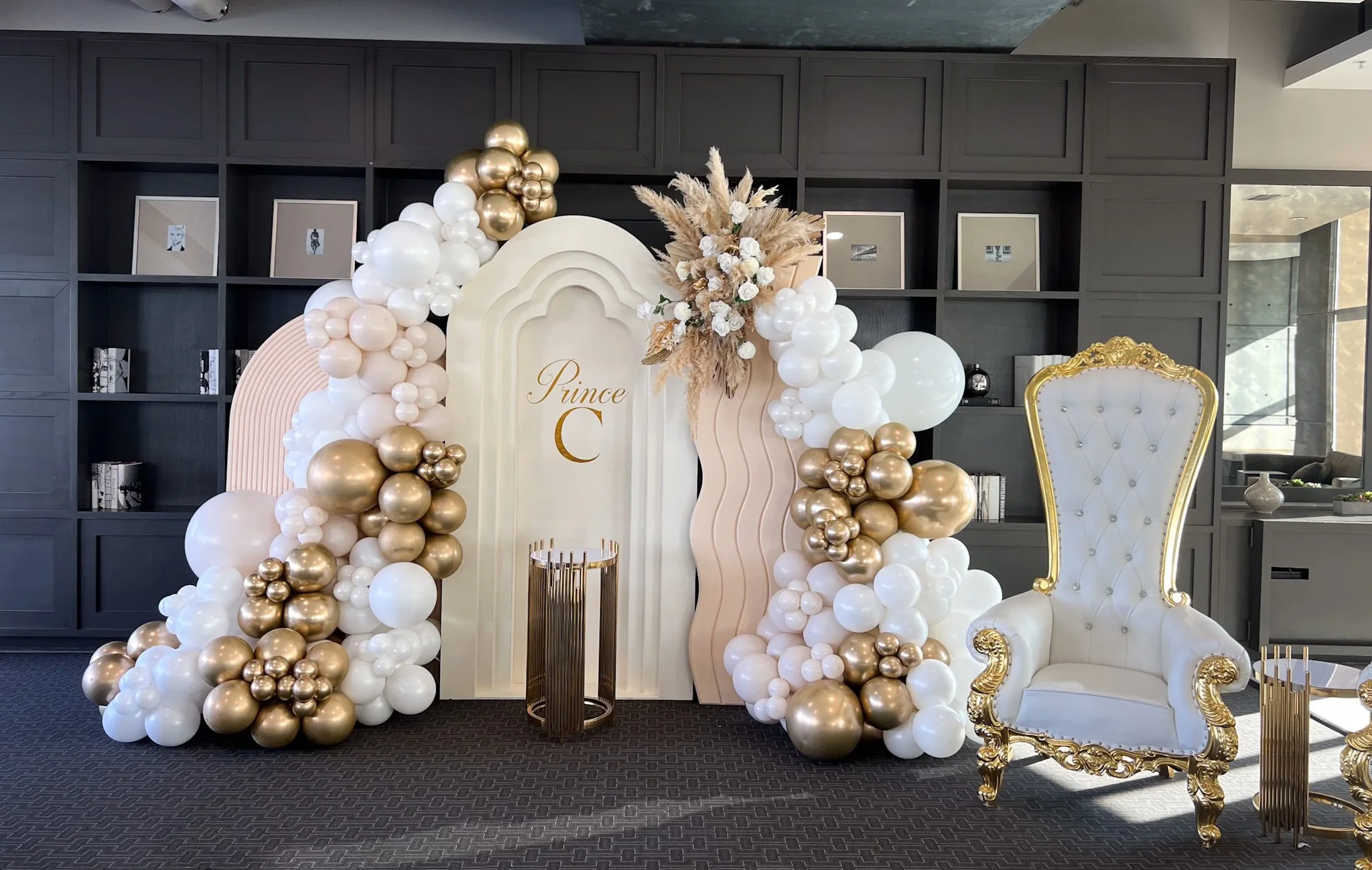 Decorative setup with a white backdrop labeled "Prince C" surrounded by balloon arrangements in gold, white, and blush pink, with a large floral arrangement at the top, and a regal white and gold throne chair to the right.