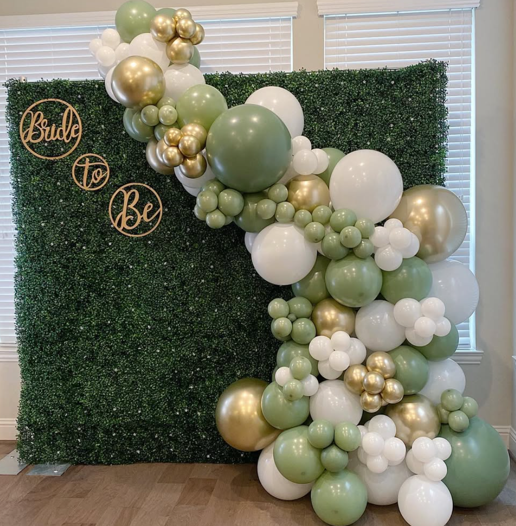 Green and white balloon arch and wall decoration with a backdrop of a green leafy hedge and a sign that says "Bride to Be" in gold script.