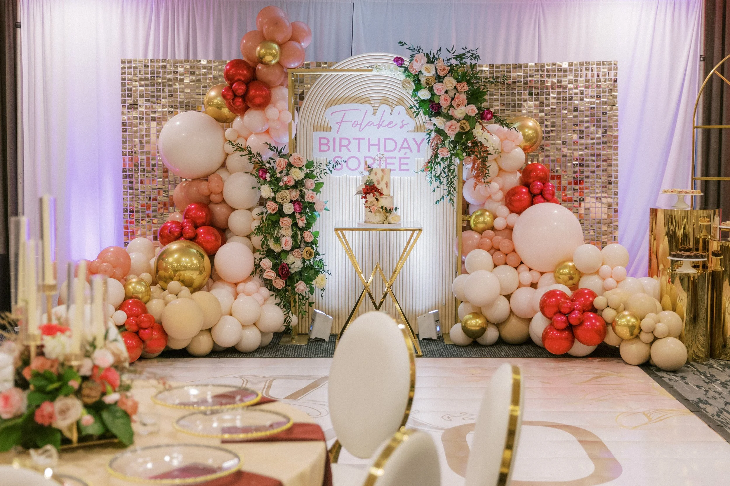 Decorative backdrop with balloons and flowers for a birthday celebration with a cake on a stand in front of an illuminated sign reading 'Folake's Birthday'.