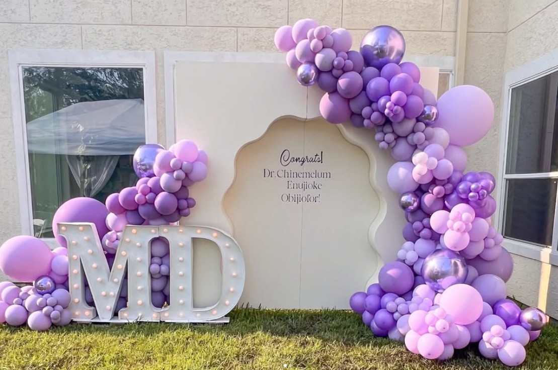 Decorative setup with a large illuminated 'MD' sign surrounded by purple, lavender, and metallic balloons arranged as an arch and cluster, with a white backdrop having a congratulatory message for Dr. Chinemelum Enuajoke Obi, and a grassy area in fro