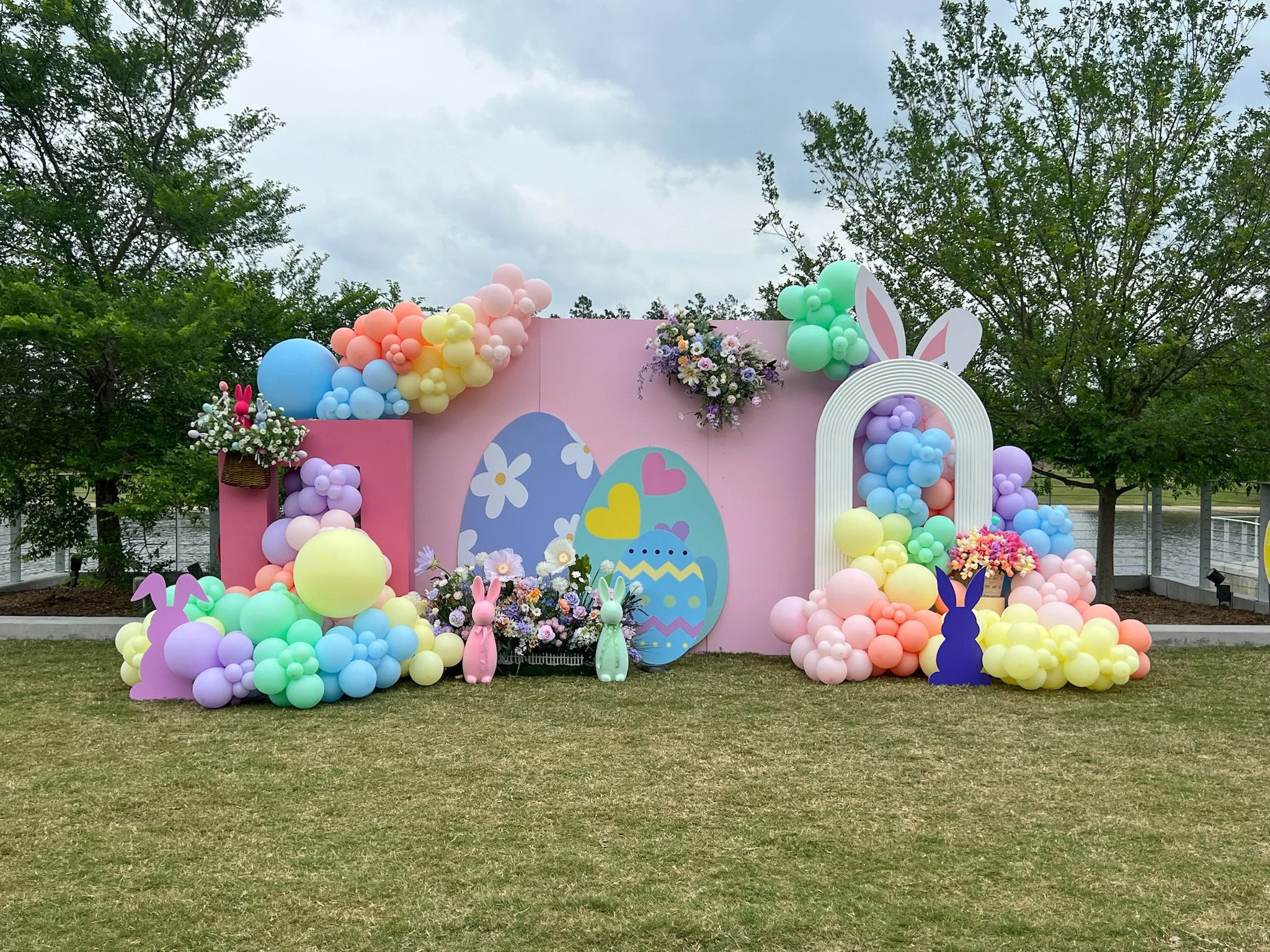 Easter-themed outdoor decoration with pastel-colored balloons, bunny cutouts, flowers, and Easter eggs against a pink backdrop with trees in the background.