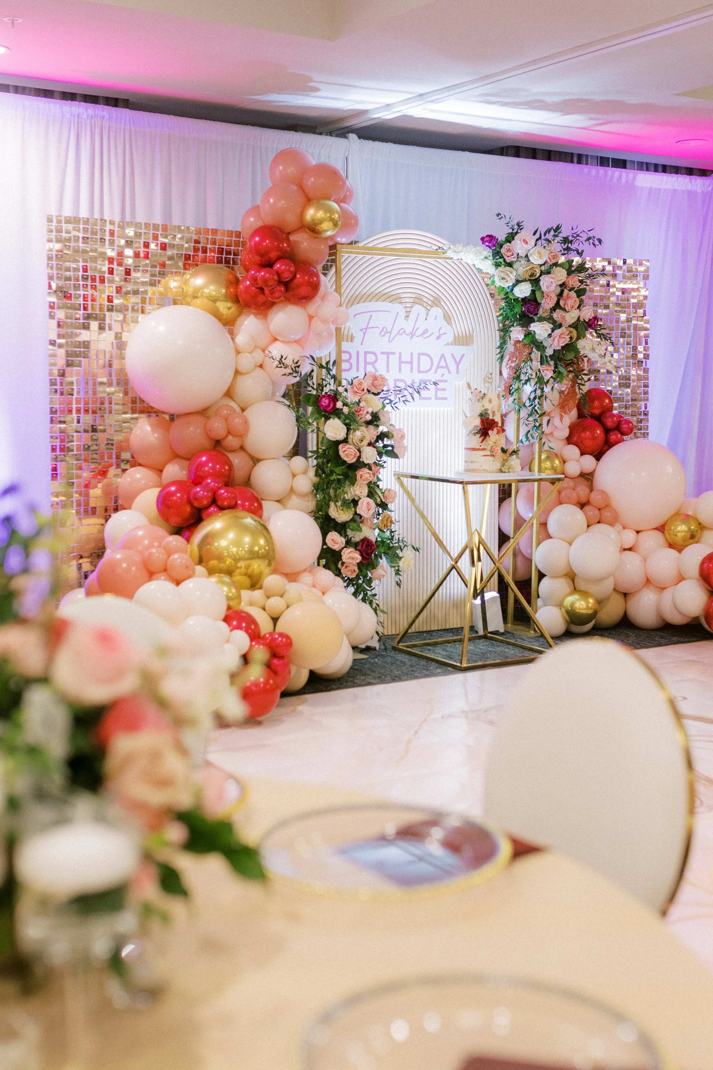 Decorative backdrop at a birthday celebration with balloons in pink, red, white, and gold, floral arrangements, and a neon sign reading "Folake's Birthday".