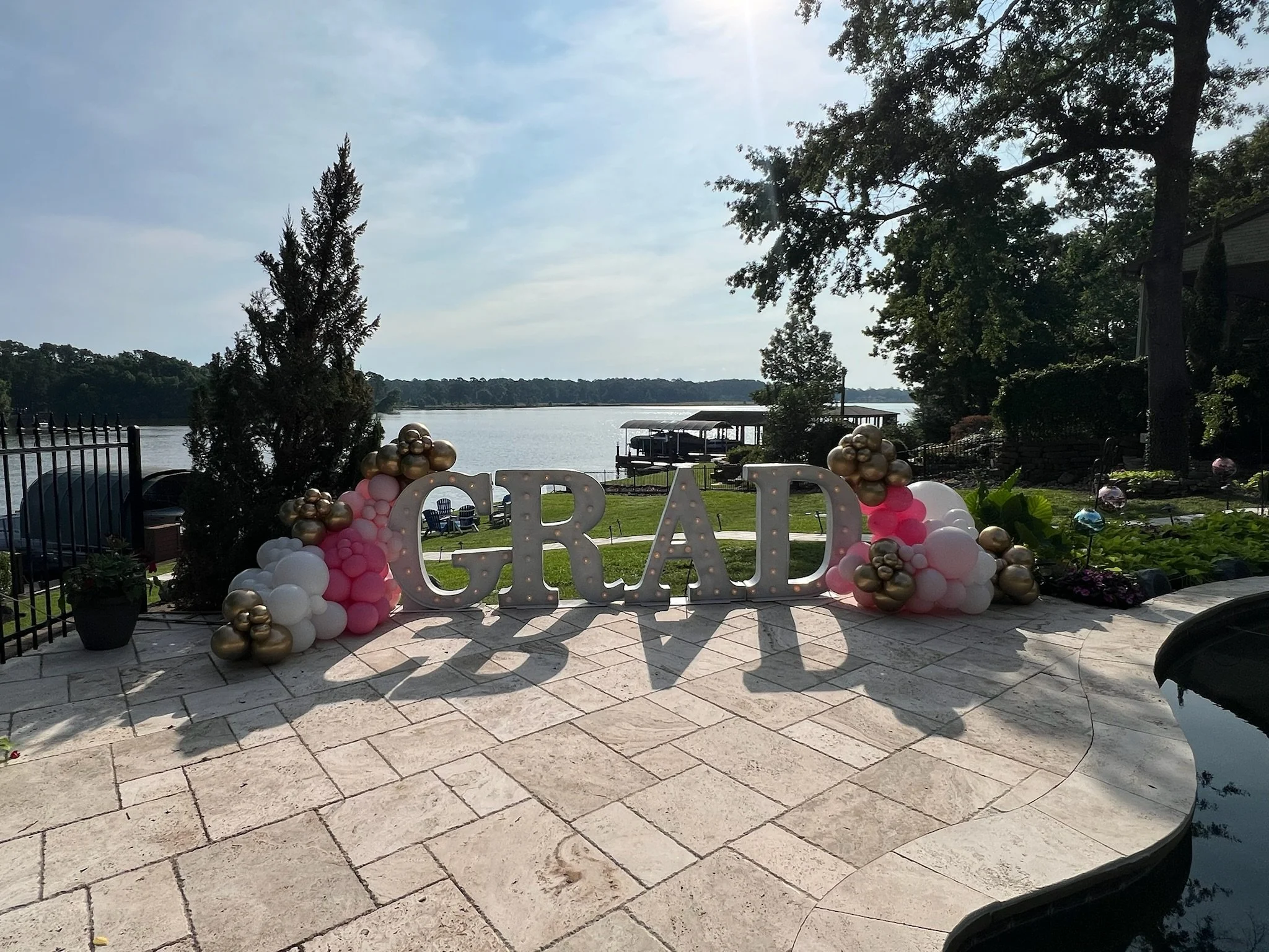 Decorative graduation celebration setup with large illuminated letters spelling "GRAD" on a patio by a lake, adorned with pink, white, and gold balloons, trees, water, and dock in the background.