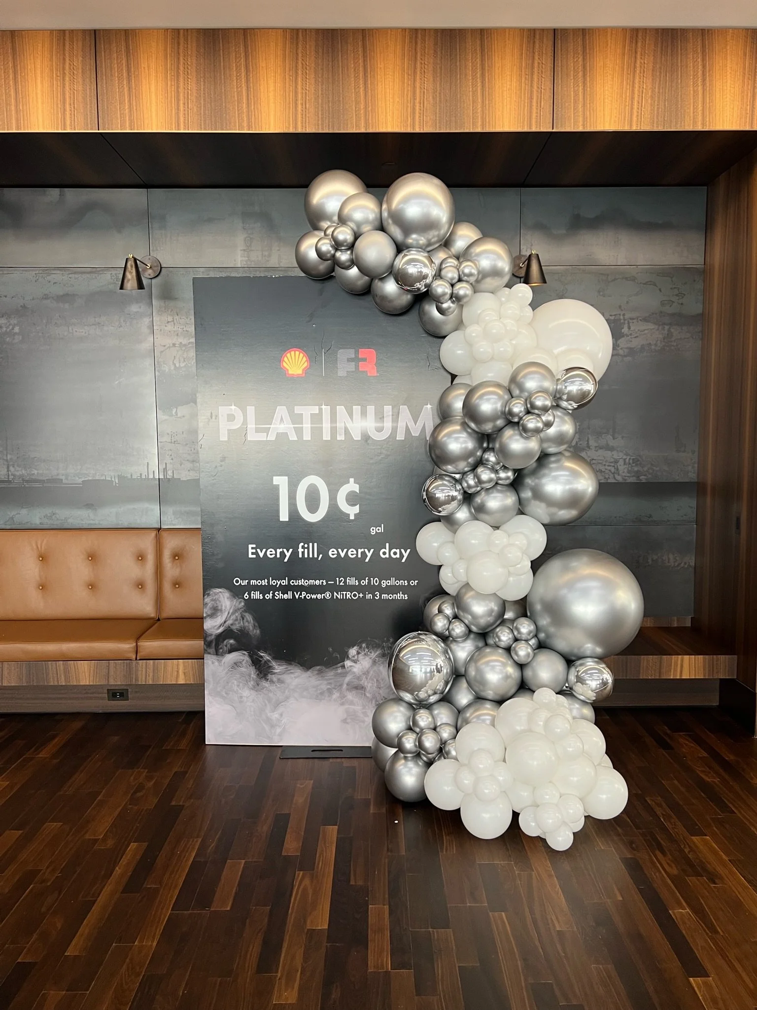Decorative display with a black sign and a large balloon arrangement of silver, white, and metallic balloons. The sign advertises fuel at 10 cents per gallon and mentions Shell V-Power Nitro. The surrounding area features wooden walls and flooring.