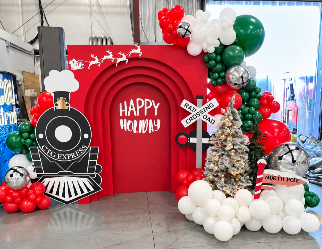 Red train-themed holiday backdrop with a CTG Express cutout, red, green and white balloons, Christmas trees, and a North Pole sign