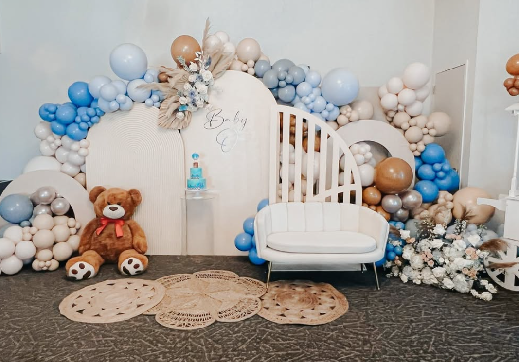 Decorative baby shower setup with pastel blue, white, and brown balloons, a teddy bear, a white loveseat, and floral arrangements.