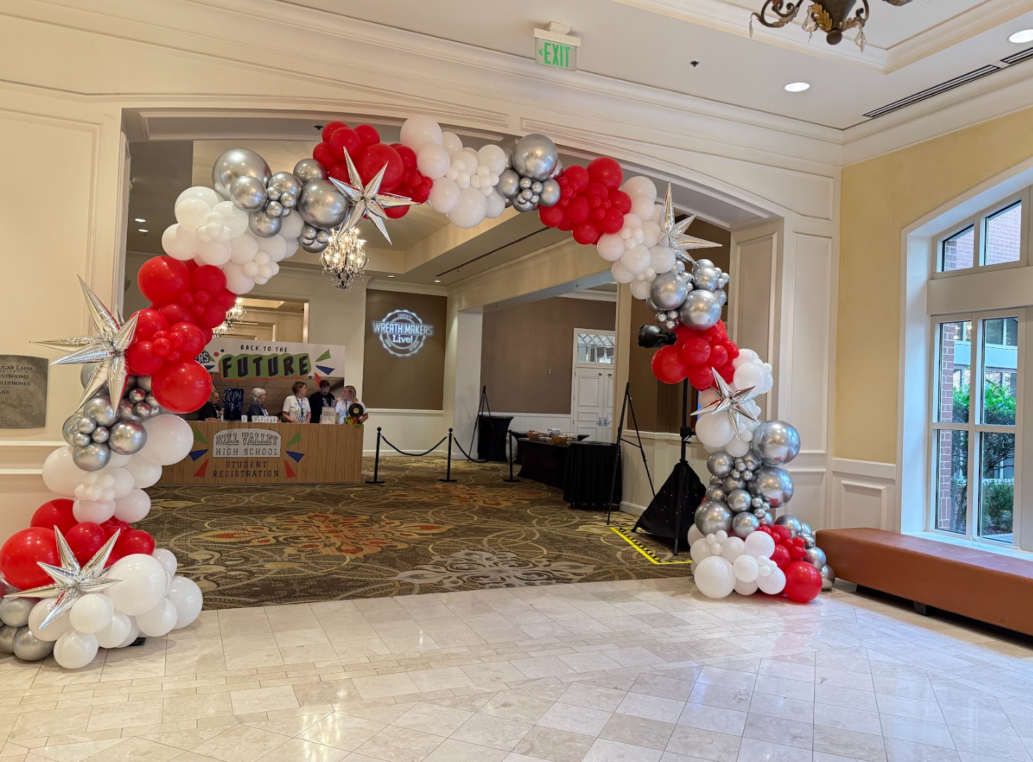 Balloon arch with red, white, silver, and black balloons, decorated with silver star-shaped balloons, forming an entrance in a lobby area. In the background, there is a registration desk for Hill Valley High School and a screen with the logo 'Wreath Makers Live!'.