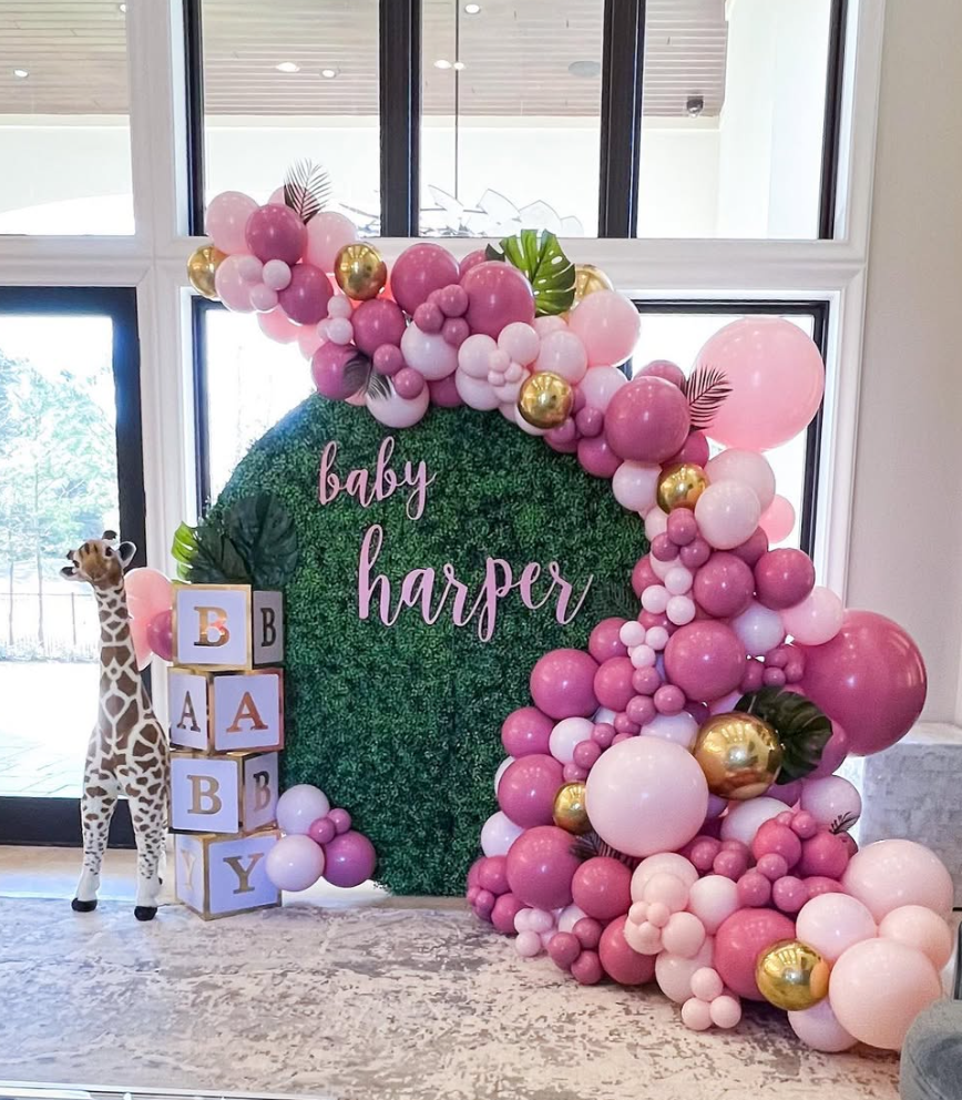 Decorative baby shower display with a green leafy backdrop that says 'baby harper' in pink script. Surrounded by pink, white, and gold balloons, with palm leaves accents. To the left, a toy giraffe stands next to stacking blocks spelling 'BABY'.