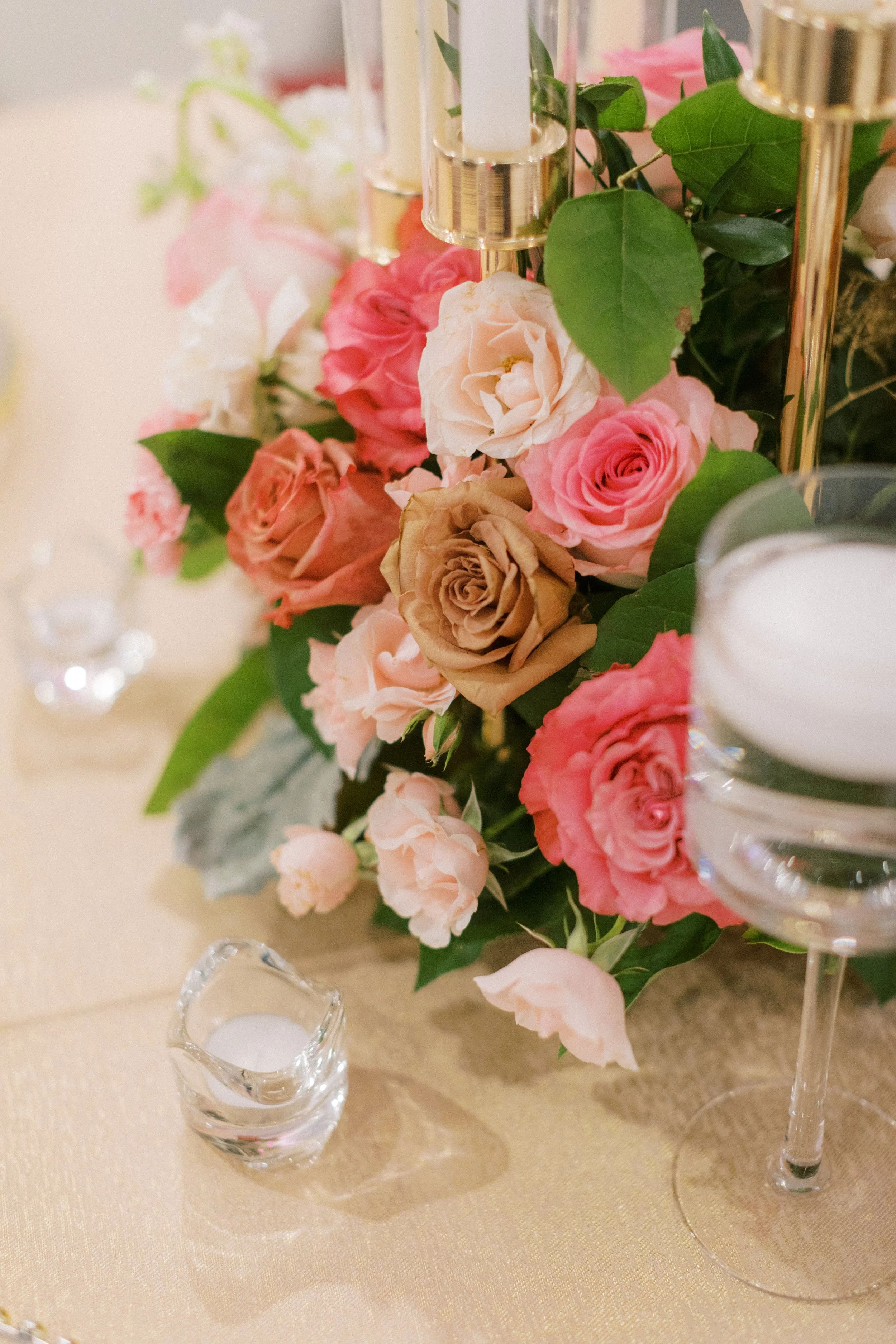 A floral centerpiece with pink, white, and beige roses, green leaves, and gold candlesticks on a beige tablecloth.