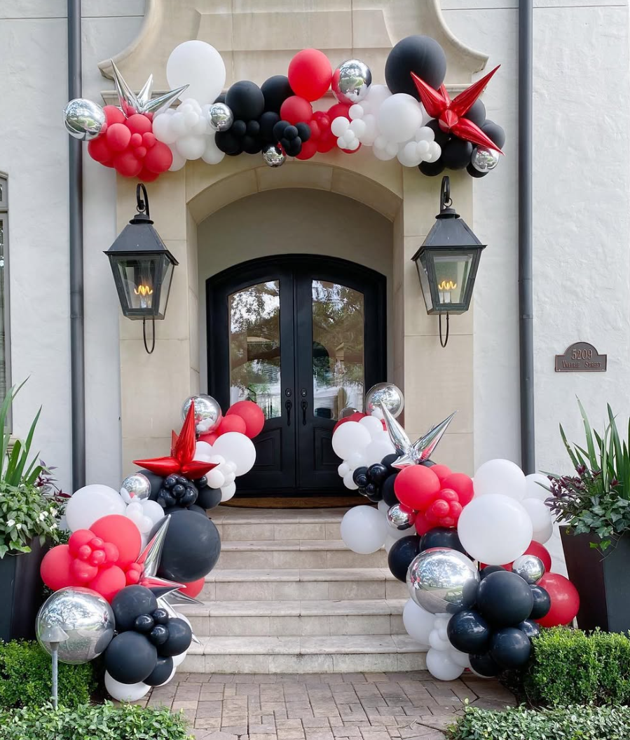 Balloon arch decoration in red, black, white, and silver surrounds the front door of a house with stairs leading up to it. There are two black lamp fixtures on either side of the door and potted plants on either side of the stairs.