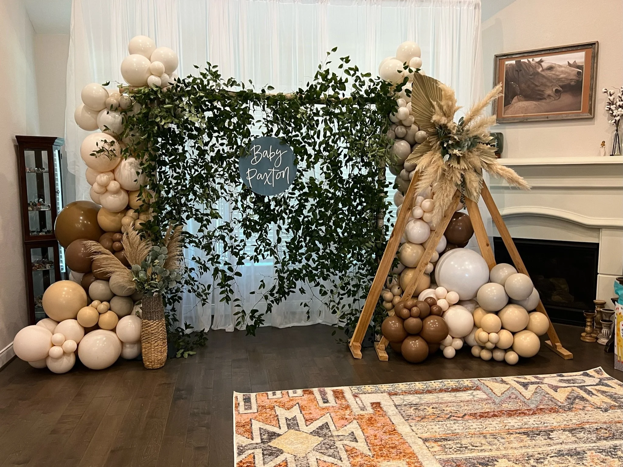 Decorative wedding or party backdrop with green foliage, beige and white balloons, and pampas grass arranged in vases, with a wooden teepee structure and a sign that reads 'Baby Paxton' in a living room.