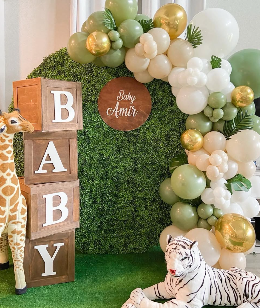 Decorations for a baby shower with a green hedge backdrop, cluster balloons in shades of green, white, and gold, wooden blocks spelling 'BABY', plush giraffe and white tiger stuffed animals, and a round wooden sign with 'Baby Amir' written on it.