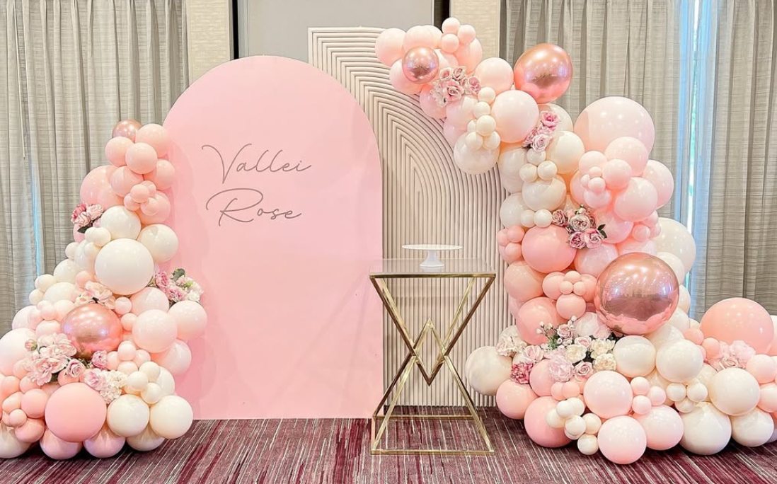 Pink and white balloon arrangement with flowers around a pink backdrop that says "Vallei Rose" and a small white cake stand on a gold table.