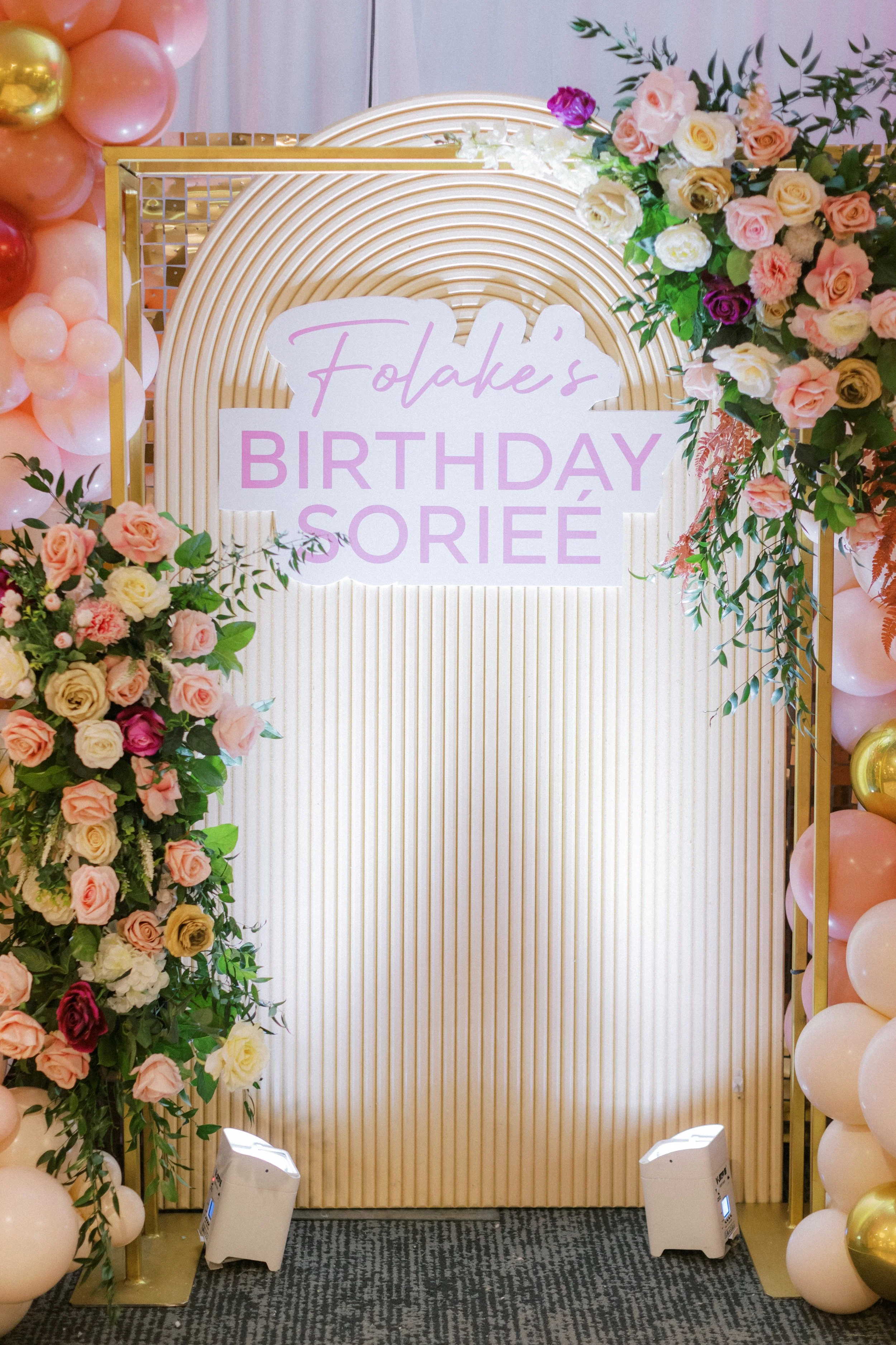 Decorative backdrop with balloons and flowers for a birthday celebration, featuring a sign that reads 'Folake's BIRTHDAY SOIRÉE'.