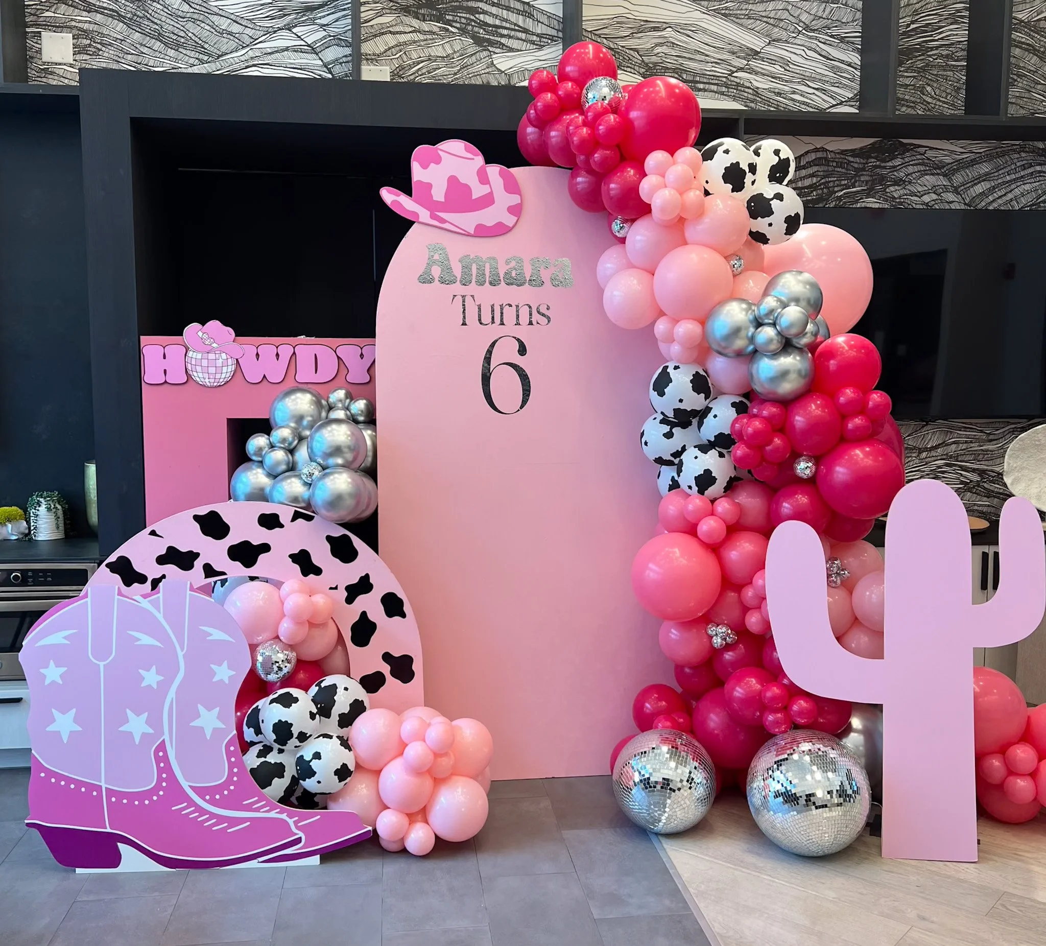 Pink and black cow-themed birthday decoration with balloons, a large pink backdrop, cowboy boots graphic, and cactus cutout, celebrating Amara's sixth birthday.