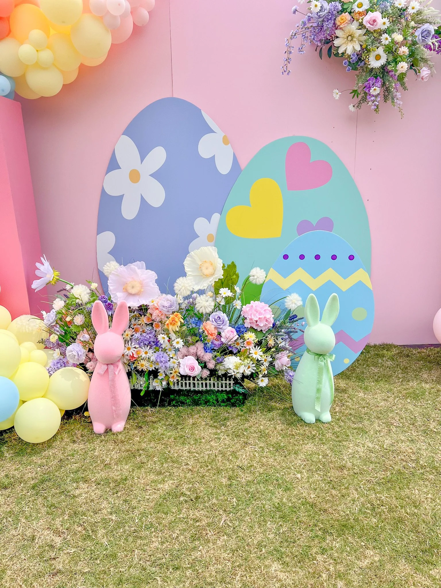 Decorative Easter display with large pastel-colored decorated Easter eggs, bunny figurines, and flowers against a pink wall with balloons and floral arrangements.
