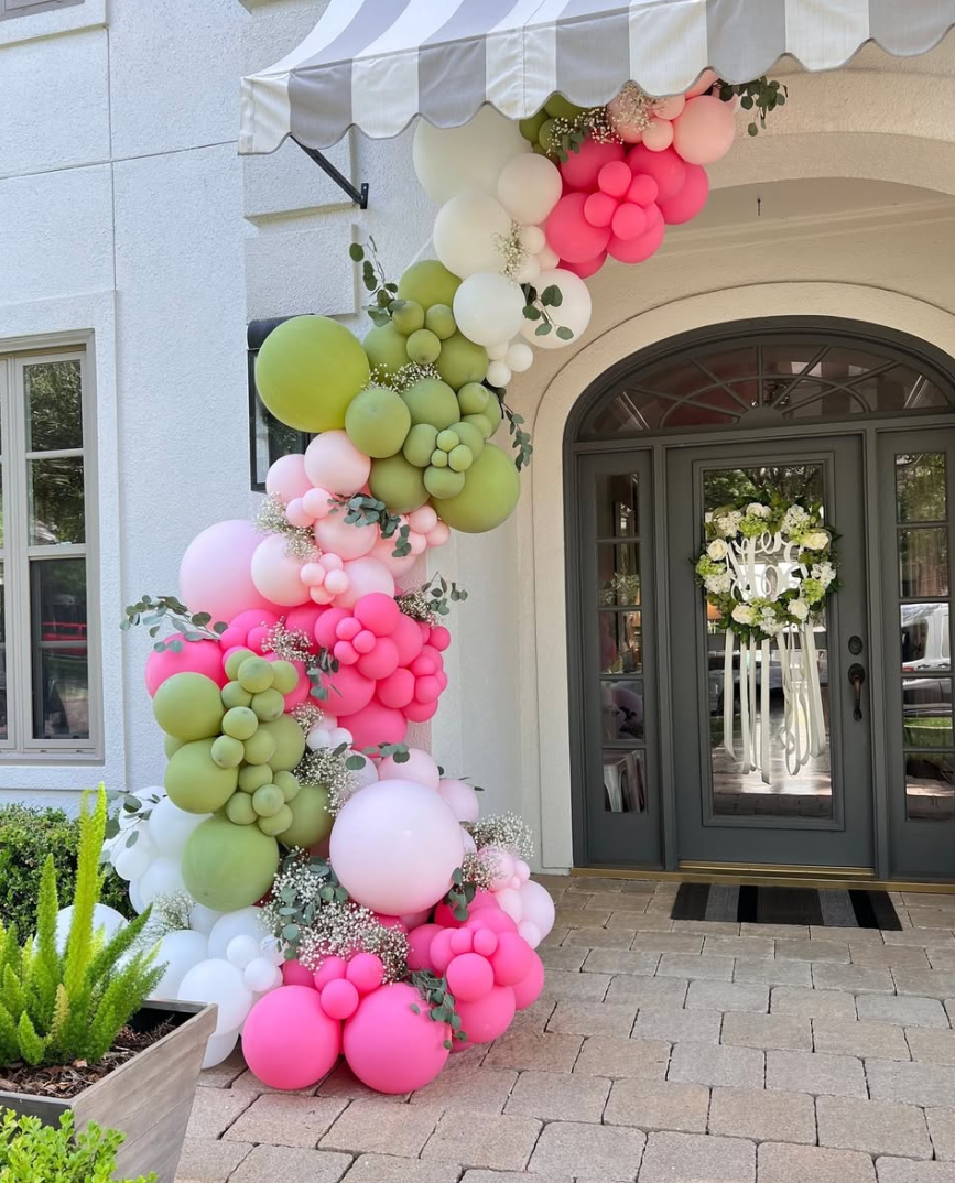 Balloon decoration arch with pink, green, and white balloons with greenery, in front of a house entrance with a floral wreath on the door.