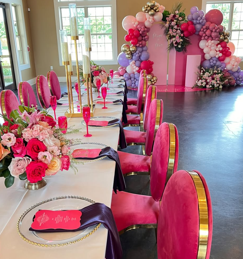 A long dining table set for an event with pink chairs, gold accents, and floral centerpieces. In the background, there is a balloon arch and floral arrangements, with large windows providing natural light.