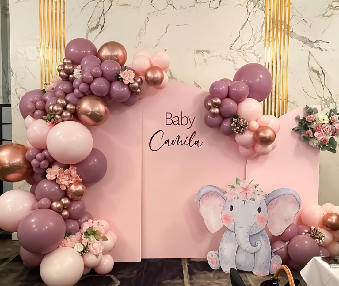 Decorative backdrop for a baby shower with pink and purple balloons, floral arrangements, and a cartoon elephant cutout, with the words "Baby Camila" written on it.