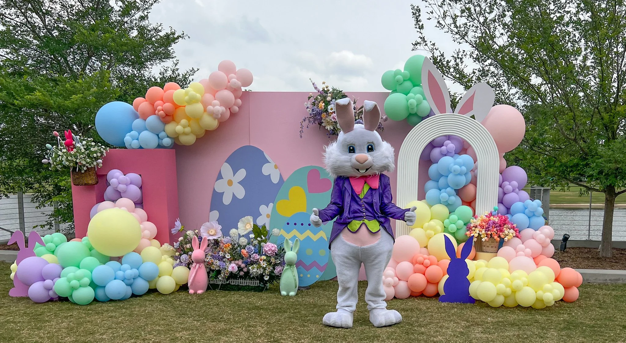 Easter-themed outdoor display with a person in a bunny costume, surrounded by colorful balloons, bunny cutouts, and floral decorations.