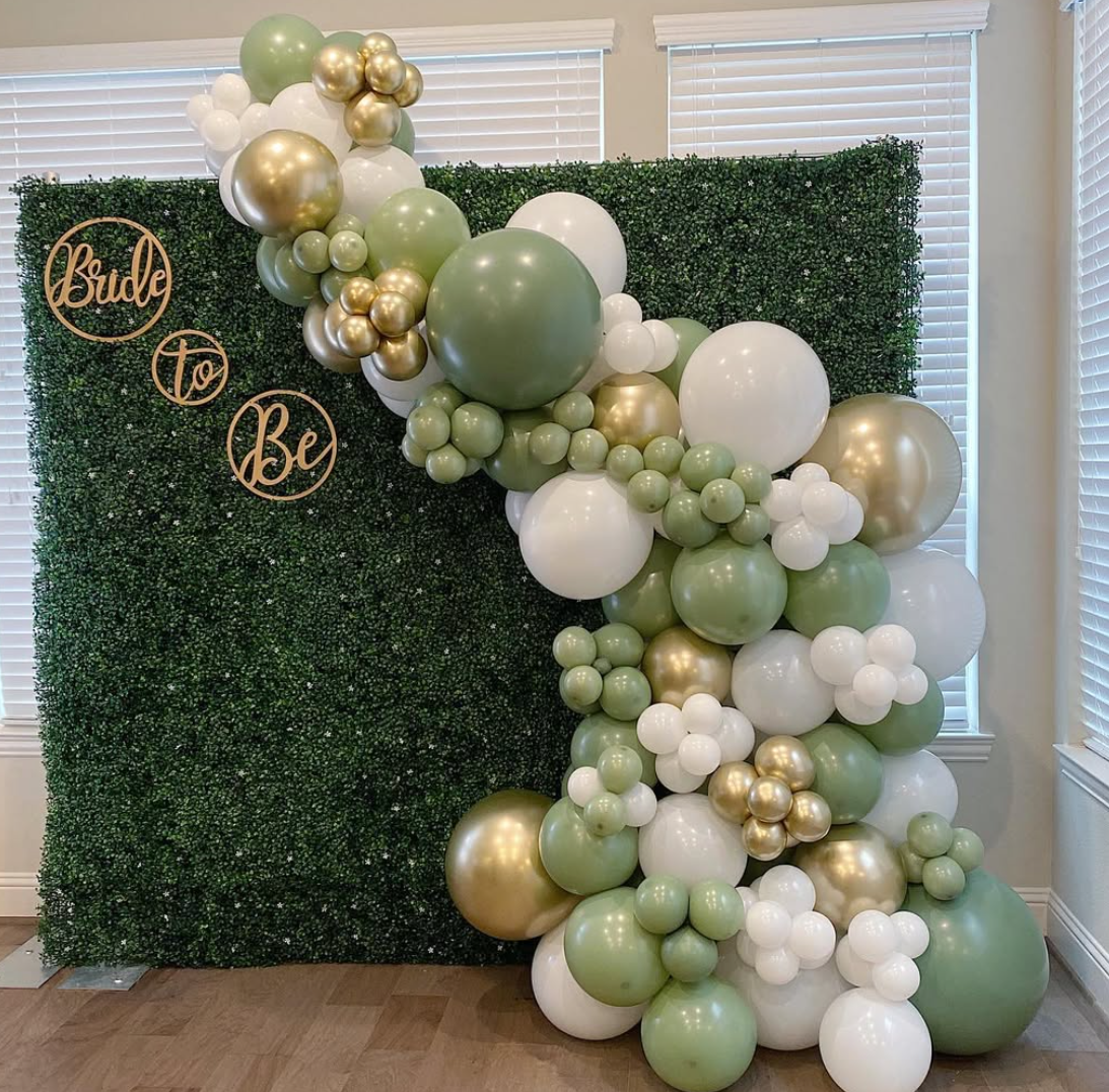 Decorative backdrop with a hedge wall and a balloon arrangement in shades of green, white, and gold, with gold signs reading "Bride" and "To Be."