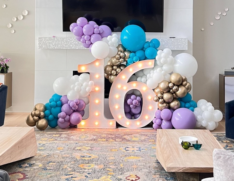 Decorative birthday display with balloons forming the number 16, including purple, white, gold, and blue balloons around large illuminated "16" numbers.