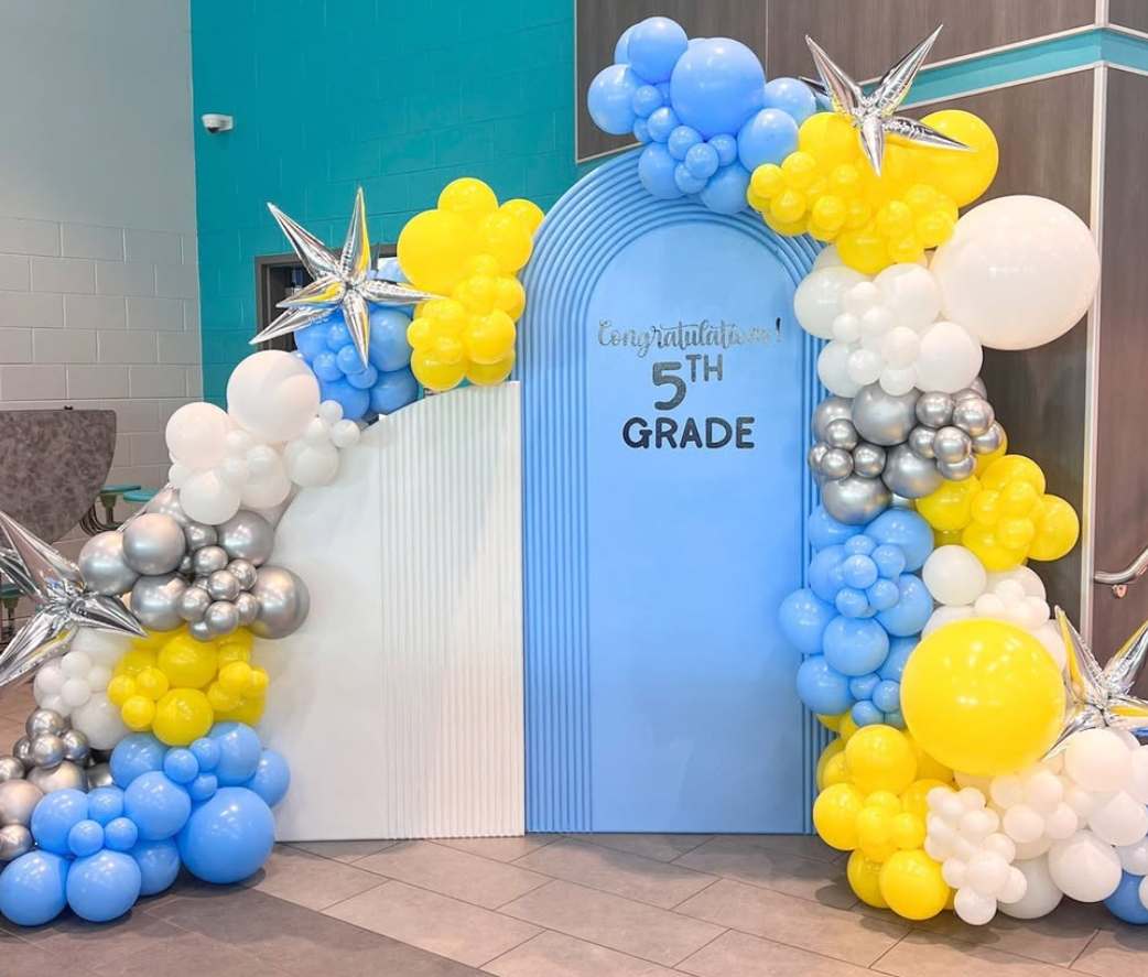 Decorative arch made of balloons in white, blue, yellow, and silver, with star-shaped balloons, surrounding a sign that reads 'Congratulations! 5th GRADE' inside a school or event venue.