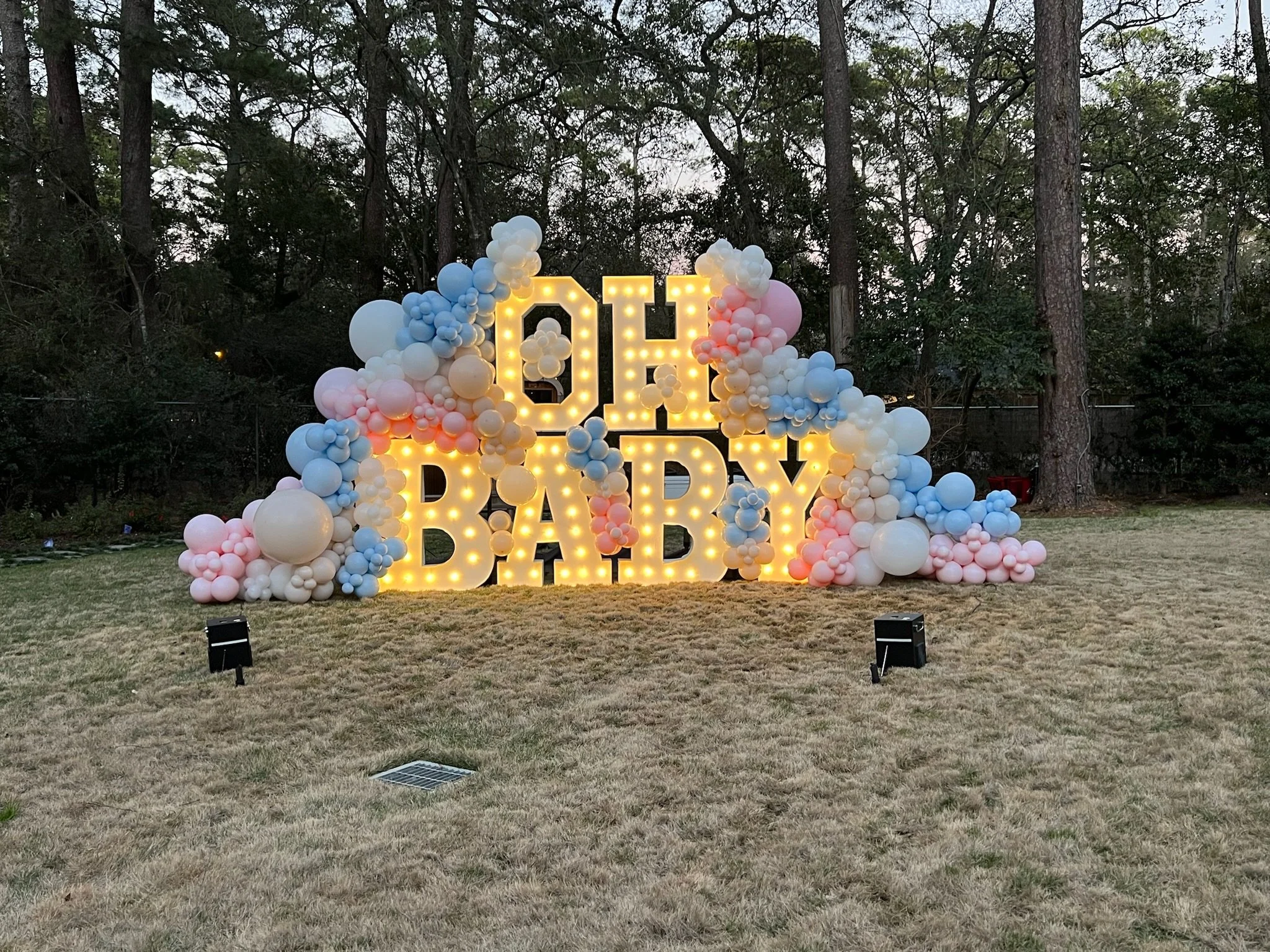 Decorative outdoor display with lit yellow letters spelling 'OH BABY' surrounded by clusters of pastel pink, blue, and white balloons against a wooded background.