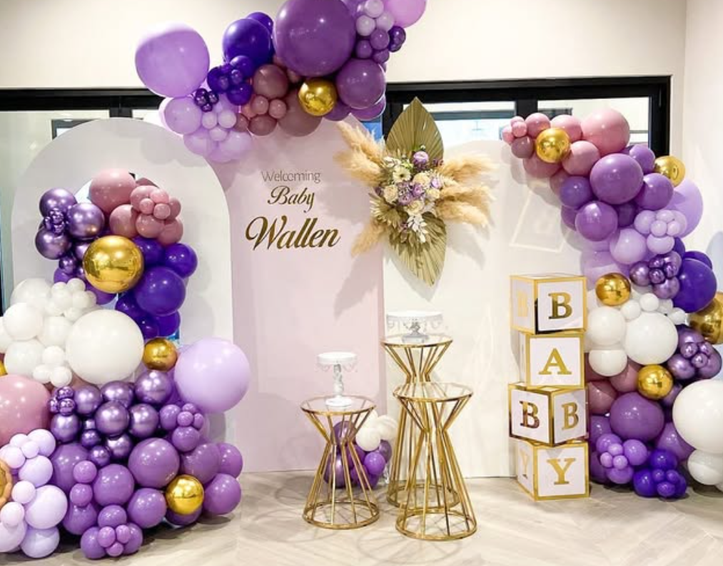 Decorative baby shower setup with purple, pink, white, and gold balloons, and a white backdrop with gold lettering that says "Welcoming Baby Wallen." There are gold geometric stands, floral arrangements with pampas grass and flowers, and blocks spell