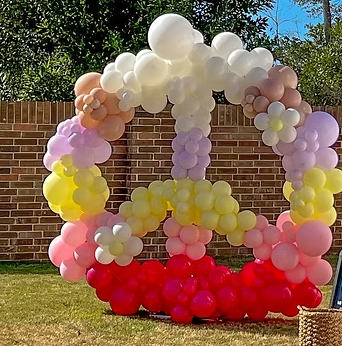 Colorful balloon arch with clouds, pink, purple, yellow, and red balloons, set up outdoors on a lawn.