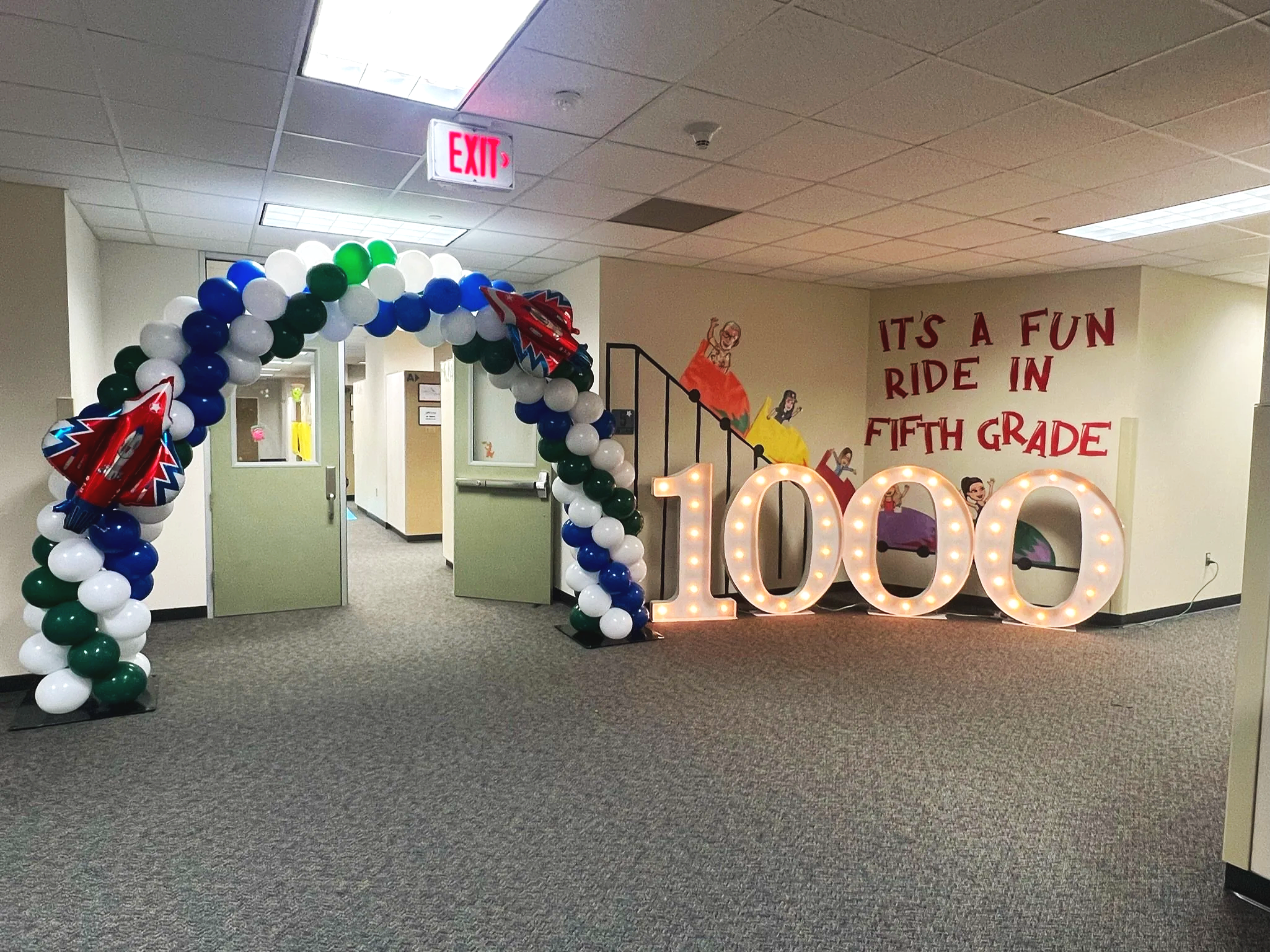 School hallway decorated with balloon arch and lit-up large number '1000' to celebrate milestone, with wall mural and banner reading 'It's a fun ride in fifth grade'.