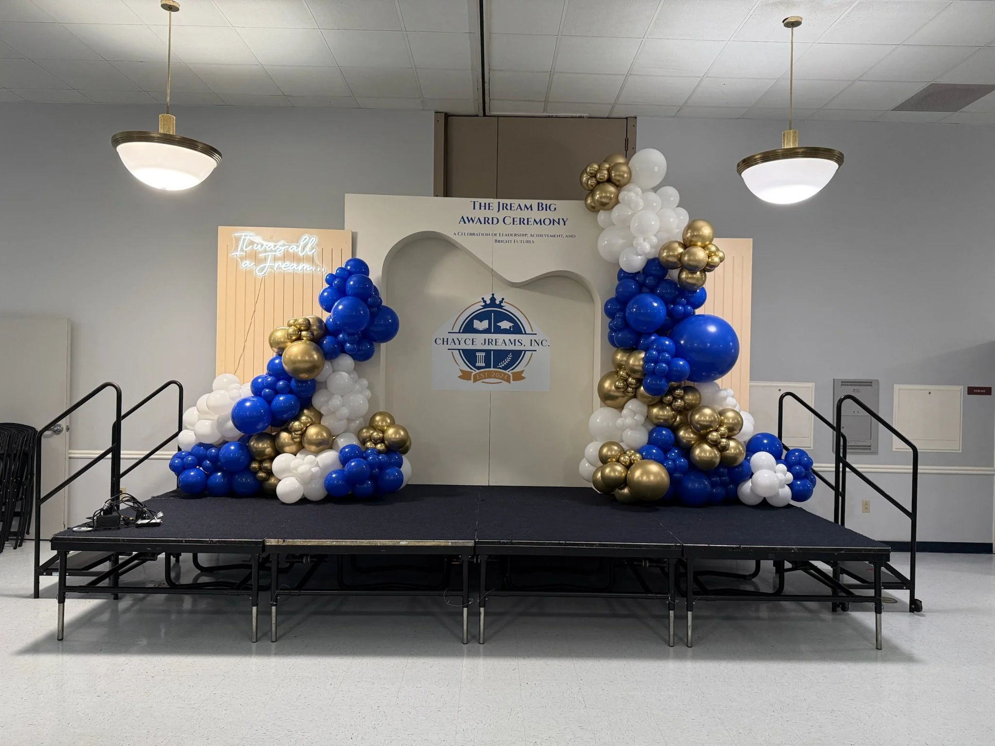 Decorated stage with blue, white, and gold balloons, backdrop with text 'The Jream Big Award Ceremony' and 'Chayce Jreams Inc.', in an event hall.