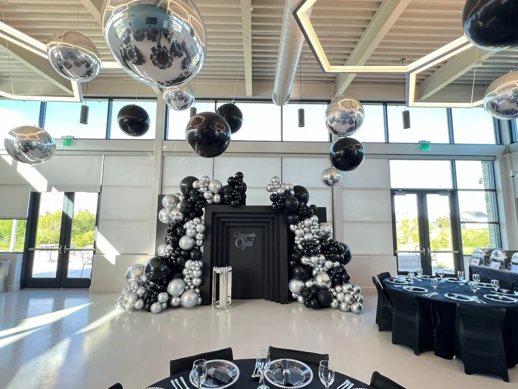 Black, silver, and chrome balloon decorations arranged in an arch and floating balloons inside a modern event space with large windows.