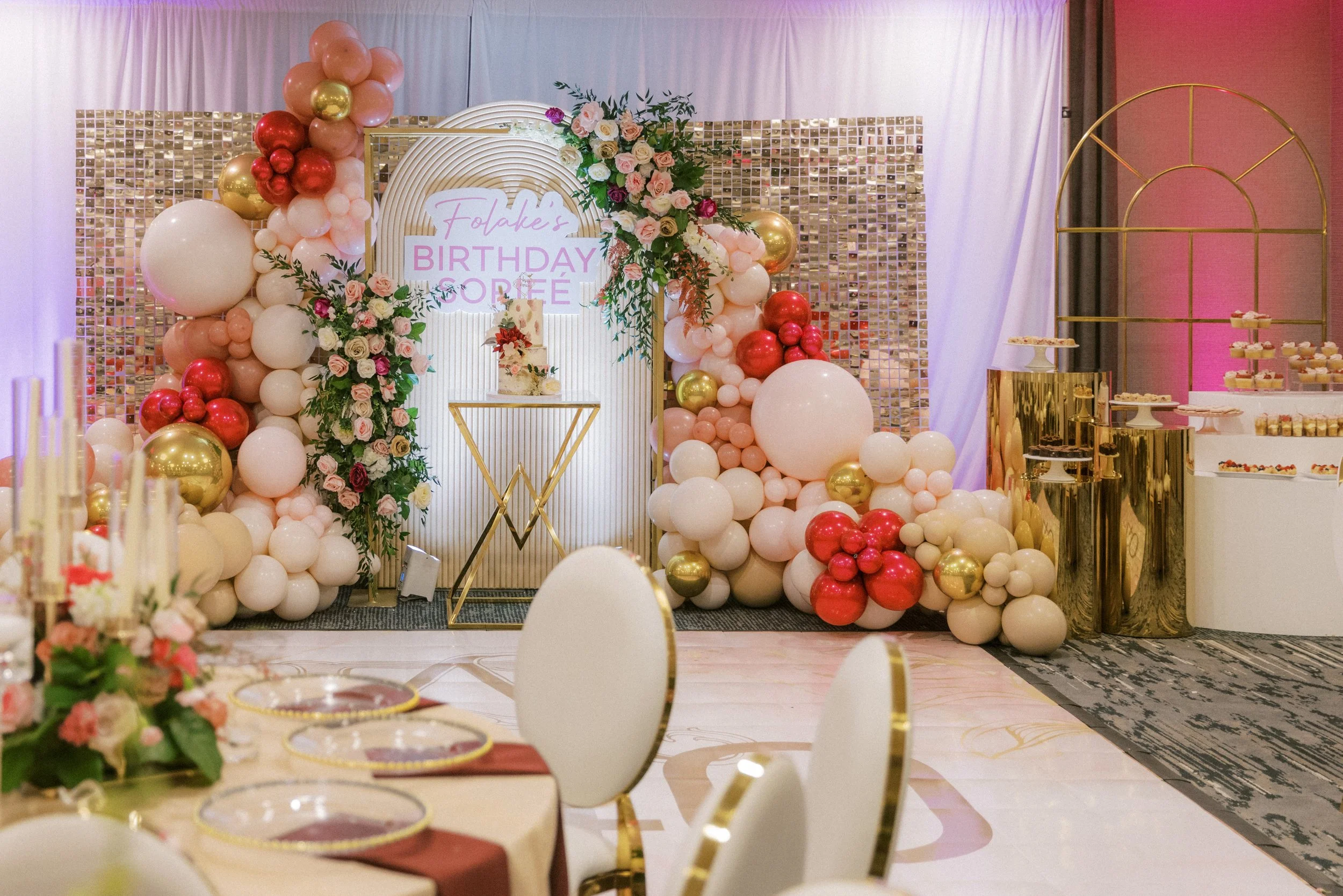 Decorative birthday party setup with balloon arch and floral arrangements, with a cake on a golden stand and a table with treats on gold stands, pink and white theme.