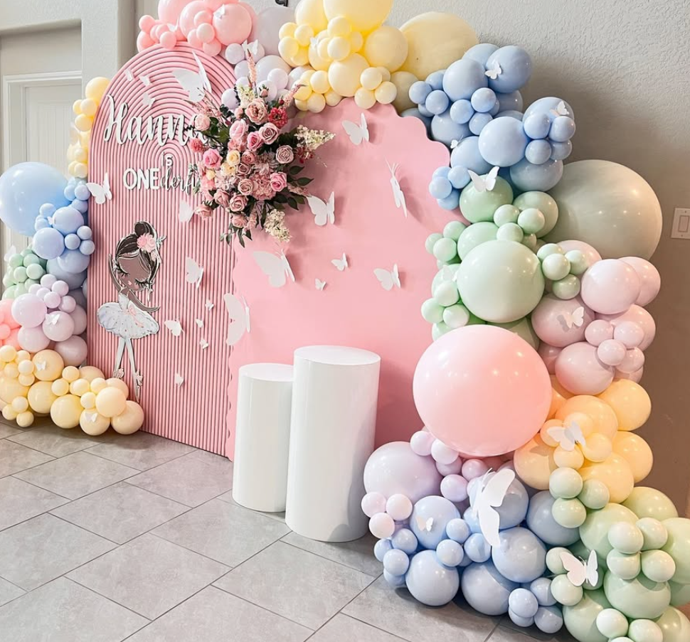Decorative birthday setup with a pink and white striped backdrop, a pink and blue balloon arch with pastel-colored balloons and white butterfly cutouts, a bouquet of pink roses and white flowers, and a drawing of a girl in a ballet tutu. The decorations include a sign that says 'Happy One-derful'.