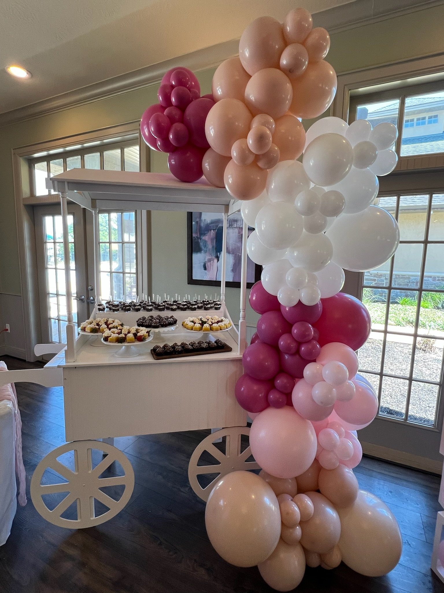 Decorative balloon arrangement with pink, peach, white, and burgundy balloons next to a white dessert cart holding cupcakes and treats in a bright room with large windows.