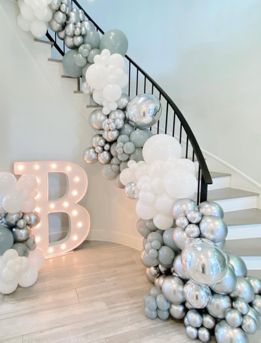 Decorative balloon arrangement in silver, white, and light gray along staircase railing with a large letter 'B' with lights illuminated underneath.