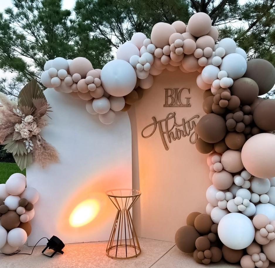 Decorative setup with balloon arch in neutral colors (white, beige, brown) and floral arrangement on the left, with a sign that reads "BIG Third" in the center, illuminated by warm lighting, outdoors with trees in the background.