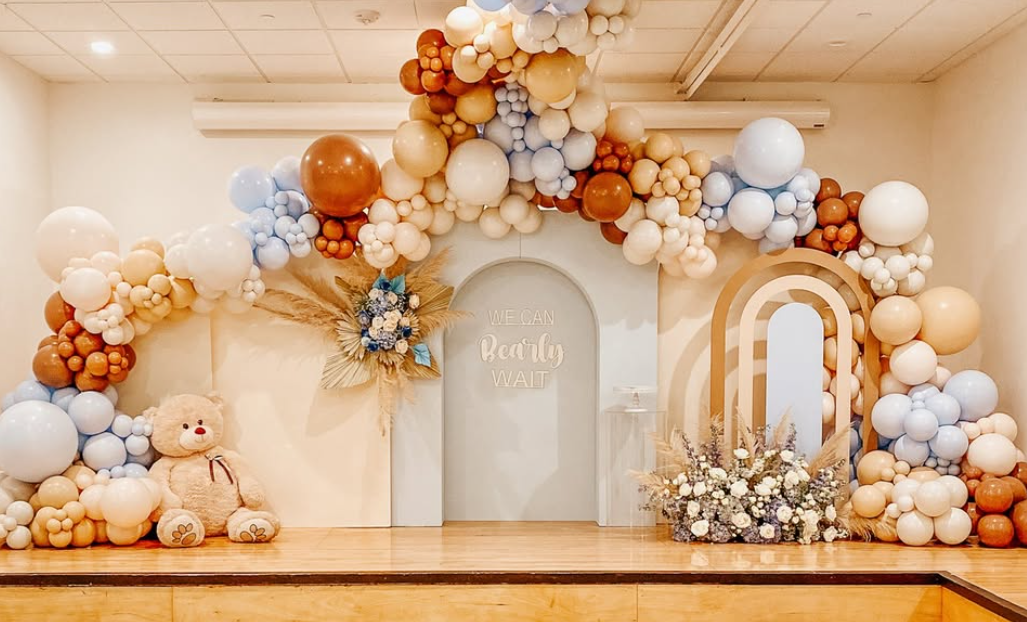 Decorative balloon arch with white, beige, brown, and light blue balloons surrounding a light blue door with a sign that says "We Can Bearly Wait." Next to the door is a teddy bear sitting on the floor, a floral arrangement, and a rainbow-shaped deco