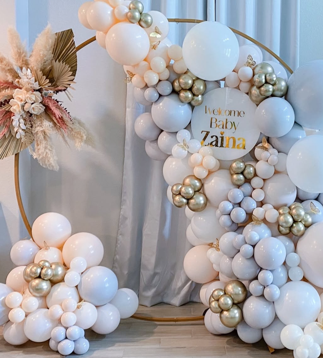 Decorative balloon arch with white, gold, and pastel pink balloons, featuring a circular sign that reads "Welcome Baby Zaina" surrounded by dried flowers and leaves on the left side.
