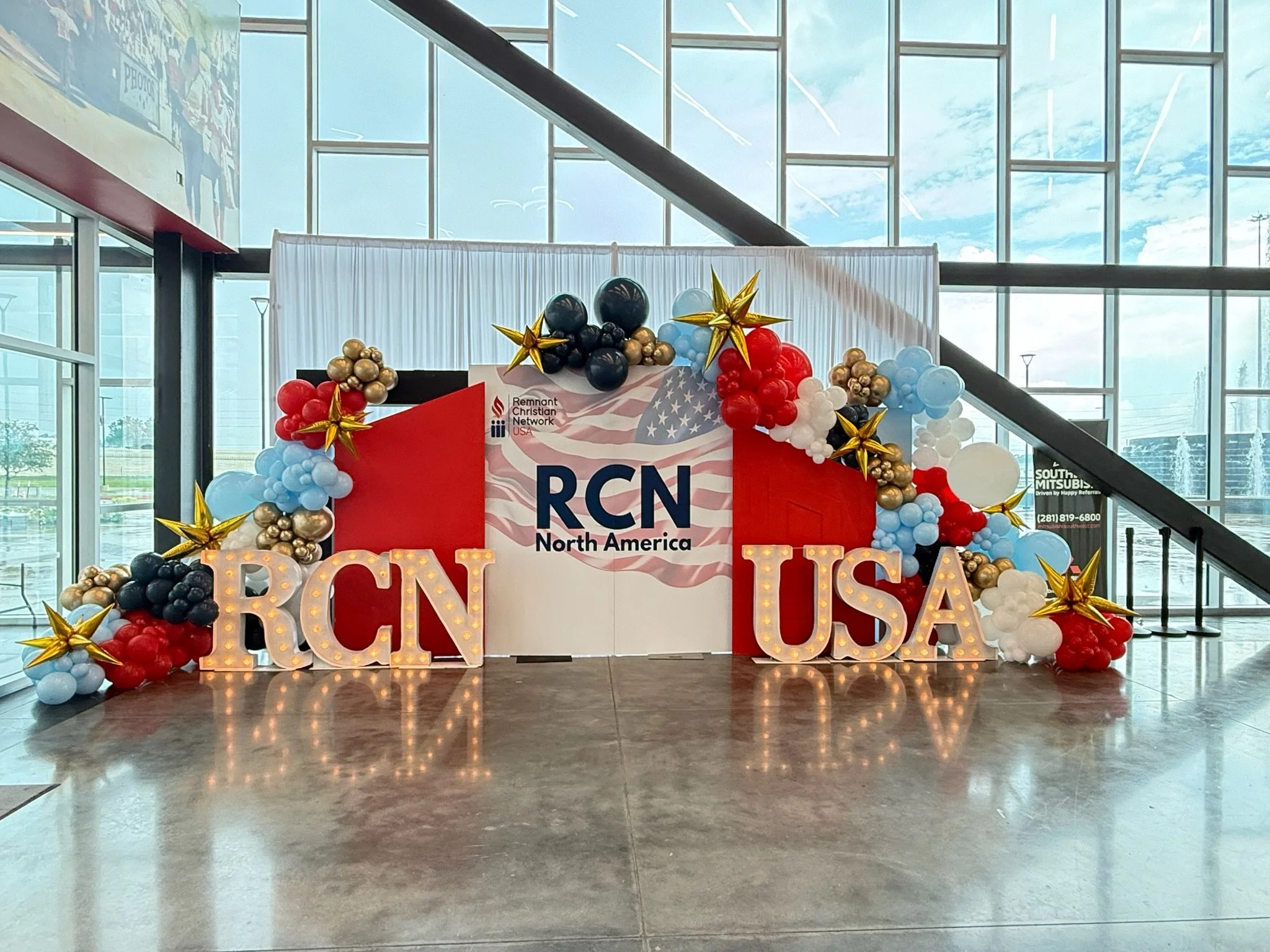 Decorative display for North America RCN event with balloons, large illuminated letters, and a backdrop featuring the American flag and RCN logo inside a modern building with glass windows and a staircase